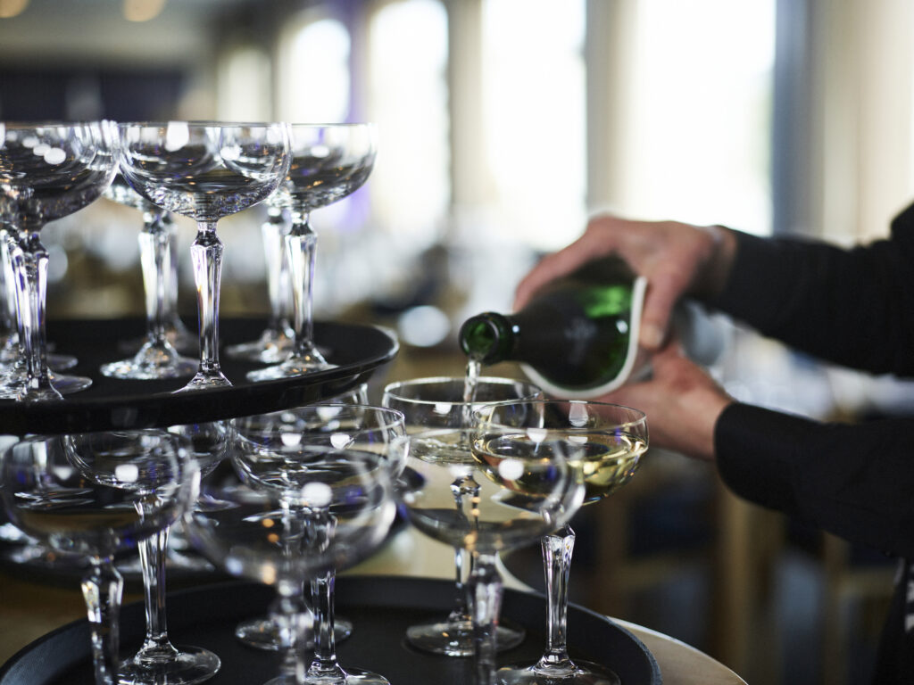 Kuohuviinilaseja tarjottimella kahdessa kerroksessa sekä tarjoilija kaatamassa kuohuviiniä laseihin.